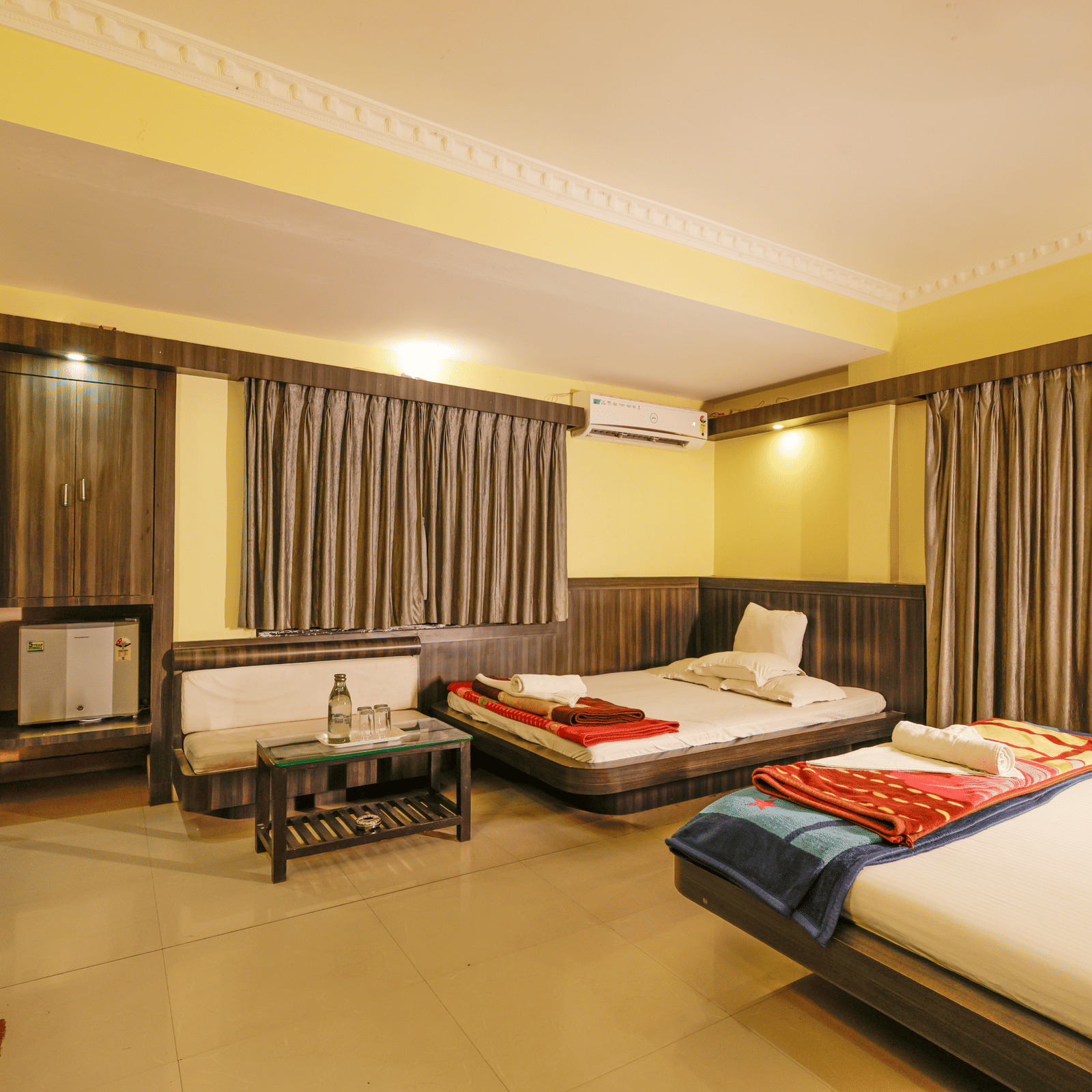 A bedroom with 2 king beds, a sofa, a coffee table, a mini fridge, curtains, and a wardrobe at Hotel Seagull Digha.